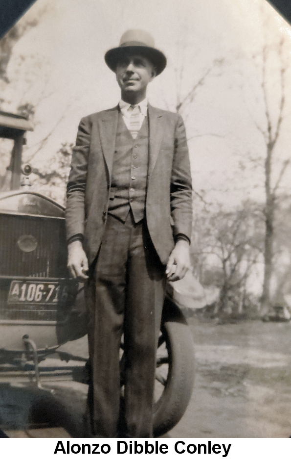 Black and white photo of a tall young man in a three-piece suit and fedora, standing in front of a 1920s-era crank-start car.